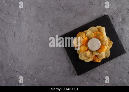 Top view of heap of potato chips with creamy white dipping sauce on black slate serving board on grey plaster table. Flavorful and Fun Chips and Dip Stock Photo