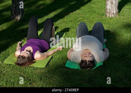 Two middle-aged women practice yoga in city park, doing exercises lying on fitness mat. Healthy lifestyle, fitness, Pilates, weight loss. overweight w Stock Photo
