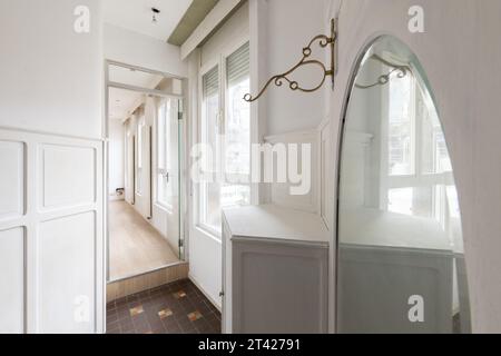 A modern interior featuring a rectangular wall mirror mounted on a white wall beside a wooden shelf and tiled floor Stock Photo