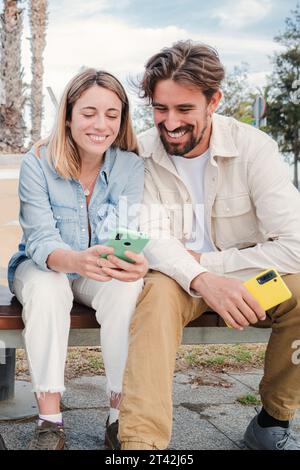 Vertical portrait of a young adult friends using their cellphones, browsing on internet enjoying free time sitting on bench outside. Happy caucasian couple having fun together with their smart phones. High quality photo Stock Photo
