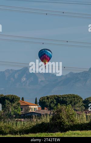 A colorful hot air balloon flies over a house and high voltage wires, with mountains in the background, Fauglia, Italy Stock Photo