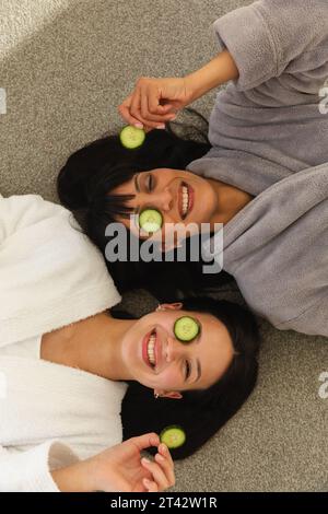 Diverse mother and daughter in bathrobes, lying on floor with cucumbers on faces at home, copy space Stock Photo