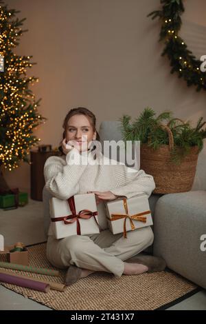 Smiling woman sitting on the floor by a Christmas tree with two wrapped gifts Stock Photo