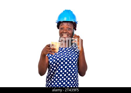 young female construction engineer wearing blue hard hat holding paper coffee cup and talking on mobile phone while smiling. Stock Photo