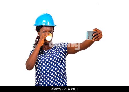 young female engineer wearing blue safety helmet and drinking paper cup of coffee while taking picture on mobile phone. Stock Photo