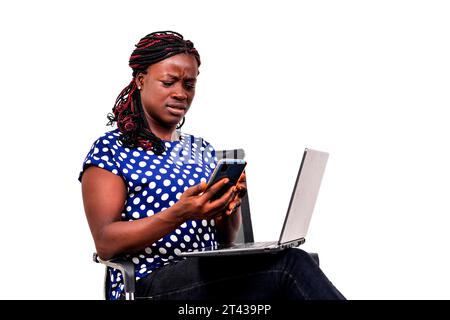 a beautiful businesswoman with braids sitting in chair at home reading message on cellphone with disgruntled look while holding laptop on thighs. Stock Photo