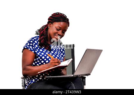 a beautiful businesswoman with the braids sitting on a chair at home working using laptop and taking notes in a notepad smiling. Stock Photo