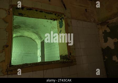 Interior of abandoned empty gloomy shabby building window with broken tiles scratched walls peeling paint adjacent to green painted brick wall Stock Photo