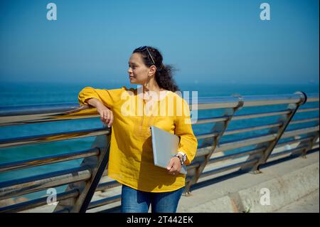 Multi-ethnic middle aged confident pensive young woman with laptop, thoughtfully looking aside the Atlantic Ocean sea. People. Lifestyle. Leisure Stock Photo