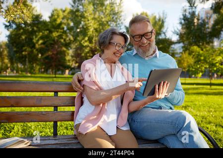 Happy cheerful senior couple using digital tablet while rest on park bench Stock Photo