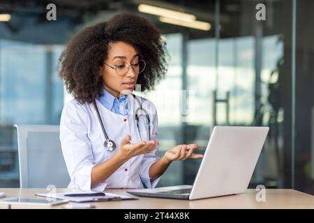 Young beautiful female doctor using laptop for video call, serious and focused woman consulting patients remotely, sitting inside clinic office, wearing white medical coat. Stock Photo