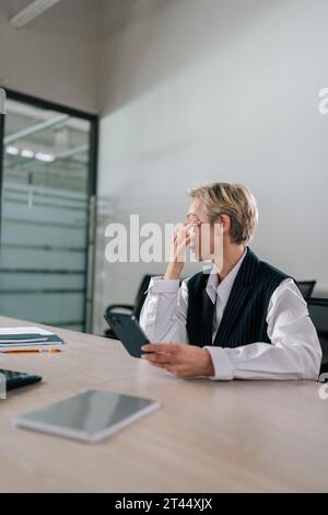 Vertical shot of exhausted middle-aged businesswoman using mobile phone at office desk, feeling discomfort from dry irritated eyes Stock Photo