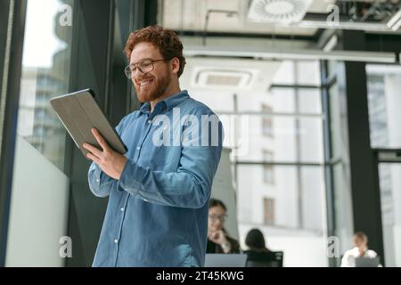 Positive bearded male manager is using digital tablet standing in modern coworking near window  Stock Photo