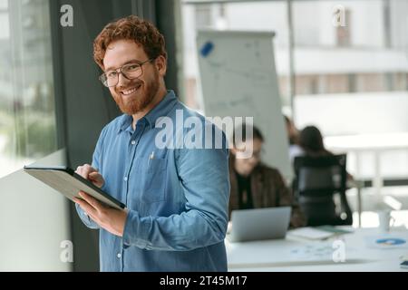 Positive bearded male manager is using digital tablet standing in modern coworking and looks camera Stock Photo