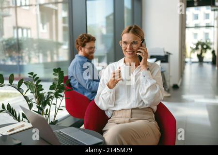 Woman freelancer talking on phone with client while working on laptop in modern coworking  Stock Photo