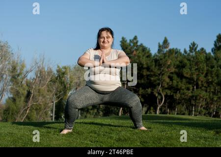 Overweight middle-aged woman doing weight loss exercises in park. Happy fat woman doing plie squats standing barefoot on grass. Healthy lifestyle, bod Stock Photo