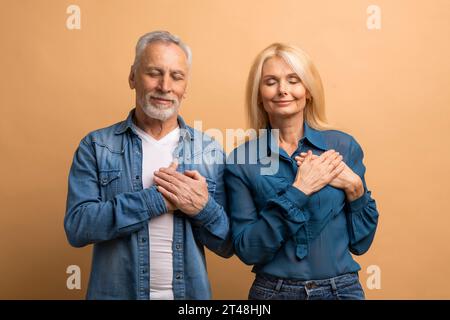 Senior man and woman placing hands on chest, beige background Stock Photo