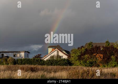 rainbow emerges from dark storm clouds above a quiet residential area, with golden grasses and autumn trees in the foreground Stock Photo