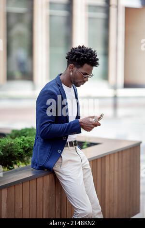 Stylish African American man in smart casual clothes, Texting on mobile phone against backdrop of city. Fashionable guy with smartphone outdoors. Stock Photo