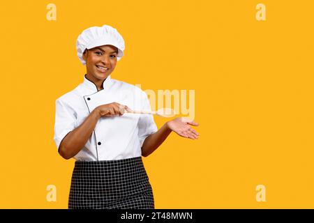 Smiling Black Chef Woman With Wooden Spatula Pointing At Her Empty Palm Stock Photo