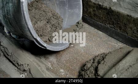 Close-up of a construction worker working on a floor. Floor screed with concrete base. A worker pours solution from a bucket onto the floor and Stock Photo
