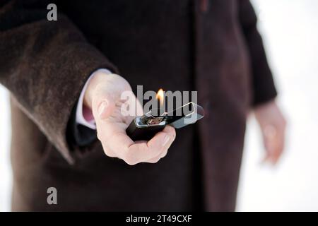 Close-up of hand with silver ring igniting a black lighter against snowy backdrop. Dark coat suggests cold setting, flame offers warmth. Stock Photo