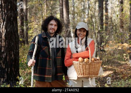Happy young intercultural couple in activewear looking at camera while smiling girl holding basket with fresh boletus mushrooms Stock Photo