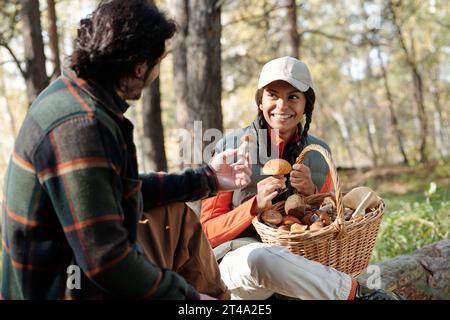 Happy young woman with basket of mushrooms looking at her husband holding fresh boletus while enjoying break in the forest Stock Photo