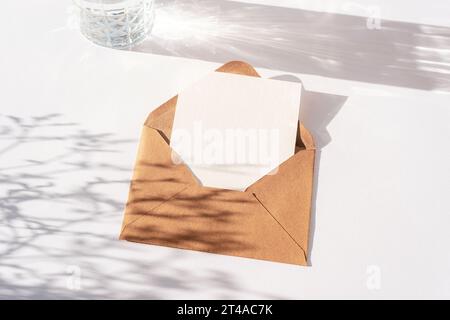 Envelope with blank card, glass of water and flowers shadow on white table. Top view, flat lay, mockup. Stock Photo