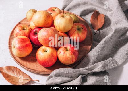 Fresh ripe apples on wooden tray, autumn leaves and grey napkin on white table. Autumn harvest, Thanksgiving concept. Top view. Stock Photo