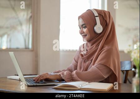 Side view image of a beautiful Asian-Muslim woman wearing a hijab and headphones is working on her laptop, having an online meeting, working remotely Stock Photo