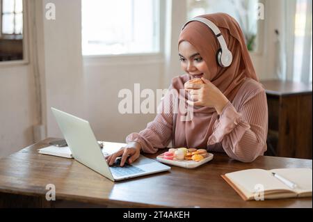 A happy young Asian-Muslim woman wearing a hijab and headphones is enjoying a doughnut while working on her laptop remotely at a coffee shop. Stock Photo