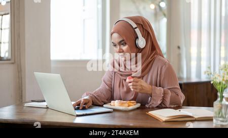 A concentrated Asian-Muslim woman wearing a hijab and headphones is eating snacks and working on her laptop while sitting in a coffee shop. Stock Photo