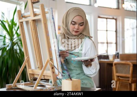 A talented Asian-Muslim female artist focuses on painting on canvas, mixing colors on a palette, working in her home studio. Stock Photo