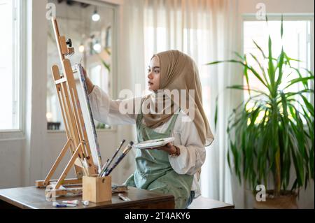 A concentrated and creative Asian-Muslim female artist creating her artwork, drawing or painting on canvas in her art studio workshop. Stock Photo