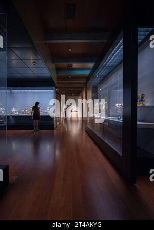 Calouste Gulbenkian Museum (Museu Calouste Gulbenkian) Lisbon, Portugal. Young woman looking at decorative tea sets in one of the galleries inside the museum.  Spotlighted at the end of the gallery is Jean-Baptiste Carpeaux’s 1872 sculpture L’Amour a la Folie (Love to madness). Stock Photo