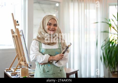 An attractive and creative young Asian-Muslim female artist in an apron and hijab is smiling at the camera and standing in her studio with painting br Stock Photo