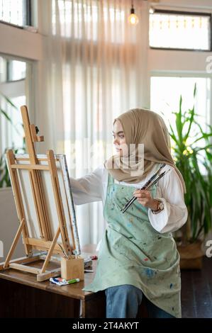 A creative and focused Asian-Muslim female artist in an apron is painting on canvas on an easel, working in her studio. Stock Photo