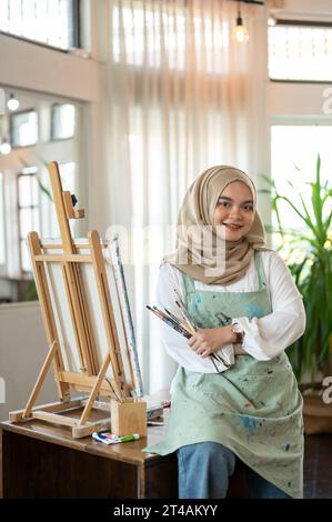 A talented and creative young Asian-Muslim female artist in an apron and hijab is smiling at the camera and standing in her studio with painting brush Stock Photo