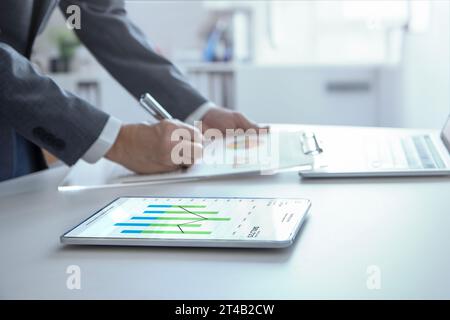 Businessman standing in the office reviewing financial annual reports with modern tablet and laptop computer for a return on investment. Stock Photo