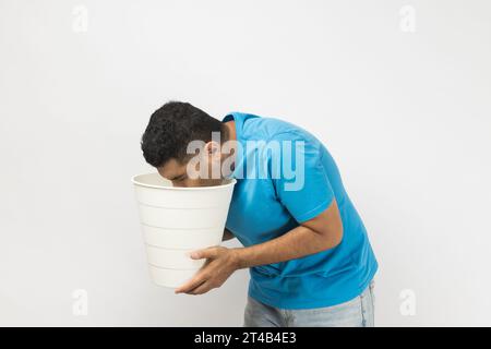 Side view portrait of sick unshaven man wearing blue T- shirt standing suffering stomachache, feels nausea and vomits, holding bin in hands. Indoor studio shot isolated on gray background. Stock Photo