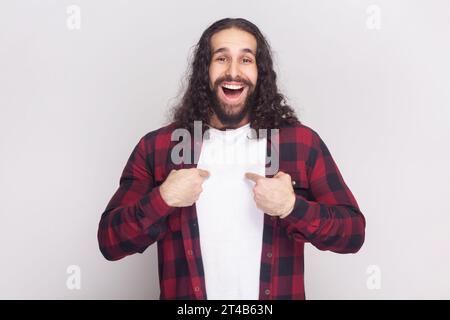 Pleased smiling bearded man with long curly hair in checkered red shirt points at himself, looks with proud happy expression boasts of his success. Indoor studio shot isolated on gray background. Stock Photo
