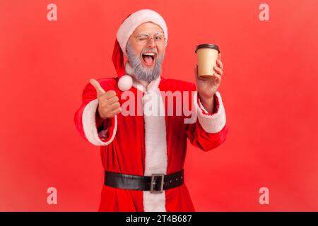 Portrait of happy elderly man with gray beard wearing santa claus costume standing with disposable paper cup with coffee, showing thumb up. Indoor studio shot isolated on red background. Stock Photo
