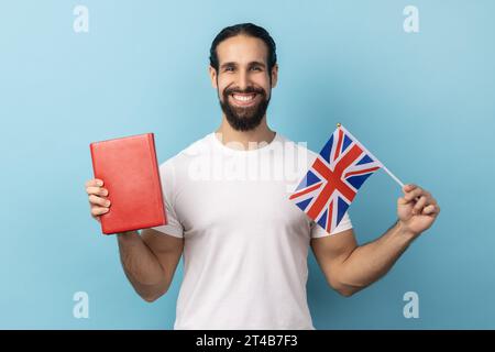 Portrait of smiling positive man with beard wearing white T-shirt holding book and Great Britain flag, education, learning English language. Indoor studio shot isolated on blue background. Stock Photo