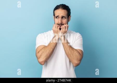 Troubles and worries. Portrait of man with beard wearing white T-shirt biting nails, terrified about problems, suffering phobia, anxiety disorder. Indoor studio shot isolated on blue background. Stock Photo