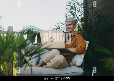 Woman in the garden working on laptop, sitting on patio chair. Businesswoman working remotely from outdoor home office, thinking about new business or Stock Photo