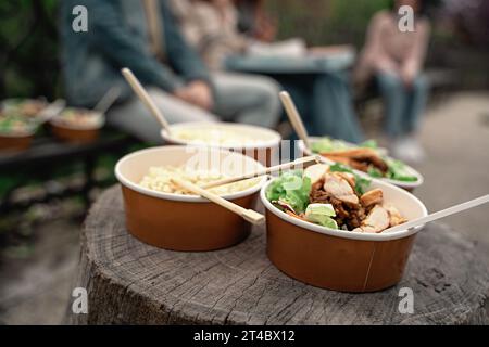 Bowls of noodles and tofu delicacies placed on a rustic wooden surface during an outdoor gathering. Blurred figures enjoy the meal, capturing a serene Stock Photo