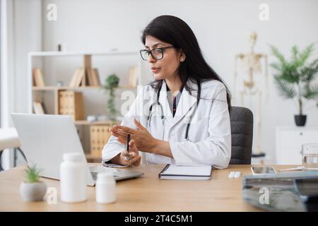 Mature indian woman in lab coat and glasses waving hello to ill patient while having conversation via computer in office interior. Family doctor using Stock Photo