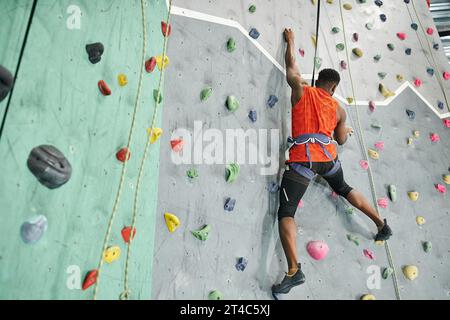 back view of young african american man in orange shirt climbing up bouldering wall with safety rope Stock Photo