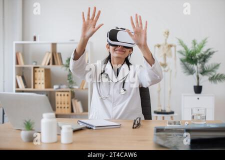 Indian nurse wearing lab coat using VR goggles while sitting at desk with modern devices on blurred background of cabinet. Female therapist preparing Stock Photo